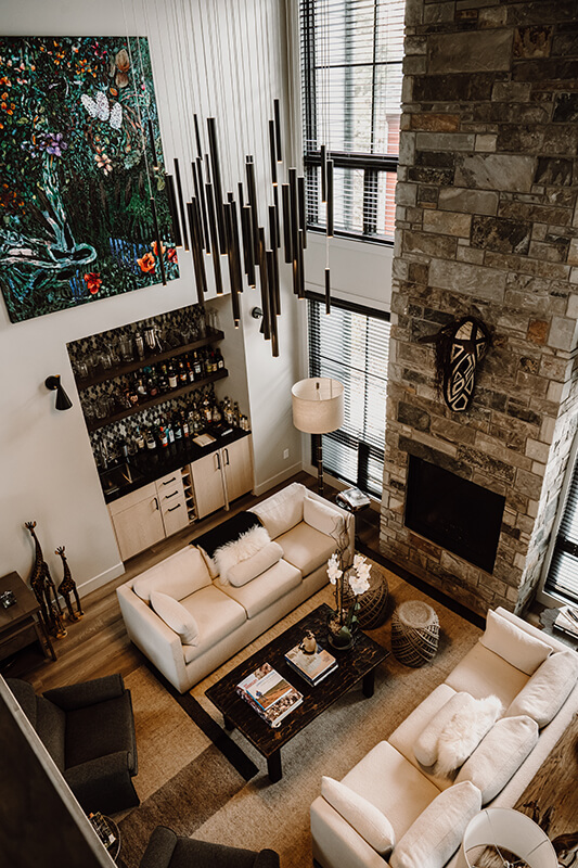 A two-story living room in Old Town Fort Collins with a mezzanine, featuring a bright and airy space painted in crisp white. The room is elegantly adorned with luxurious global finishes, including rich textures and decorative accents that blend various cultural influences. Large windows flood the space with natural light, highlighting the sophisticated design and creating a warm, inviting atmosphere.